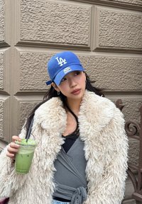Młoda kobieta w niebieskiej czapce z napisem LA i kremowym futrzanym płaszczu, trzymająca zielony mrożony napój, opierająca się o teksturowaną beżową ścianę.