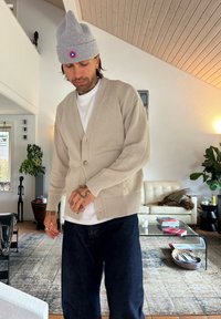 Beige gebreide cardigan met geribbelde manchetten en zoom, gelaagd over een wit T-shirt, gecombineerd met donkerblauwe jeans. Grijze beanie met een kleurrijke badge.