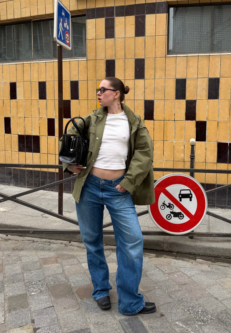 Jeune femme en vêtements décontractés se tient près d'un panneau de signalisation interdisant les voitures et les motos, avec un mur carrelé jaune et marron en arrière-plan.