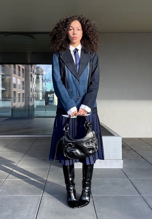 Frau mit lockigem Haar, die einen Color-Blocking-Blazer, ein weißes Hemd, eine Krawatte, einen marineblauen Faltenrock und schwarze kniehohe Stiefel trägt, eine schwarze Handtasche hält und im Freien steht.