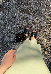 Black slide sandals featuring thick straps, paired with a green scalloped dress and a small black velvet bag adorned with gold accents. The ground surface is pebbled.