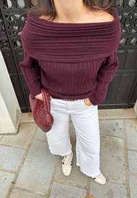 Burgundský žebrovaný svetr s odhalenými rameny, bílé široké kalhoty se třepícím se lemem a červená pletená kabelka; tenisky s krémovým základem a bordó akcenty.