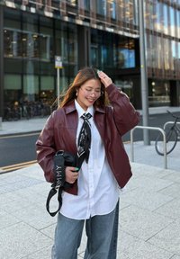 Barna bőr bombázó dzseki, fehér ing, fekete mintás sál, laza kék farmernadrág és fekete táska fehér felirattal. Tiszta városi háttér.