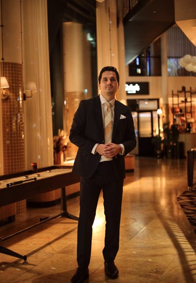 Hombre con traje oscuro, corbata de color claro y pañuelo blanco en el bolsillo, de pie en un vestíbulo moderno con azulejos en el suelo y una iluminación cálida.