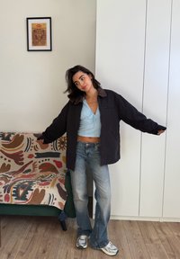 Donna che indossa una giacca nera oversize, un top corto azzurro, jeans blu e sneakers, in piedi vicino a una coperta decorata su un divano verde.