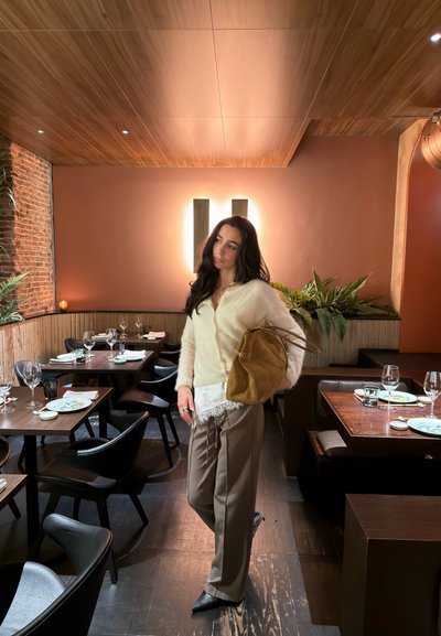 Mujer de cabello largo y oscuro, vestida con suéter color crema y pantalones anchos, sosteniendo una bolsa marrón, de pie en un restaurante con iluminación cálida, decoración de madera y mesas preparadas.