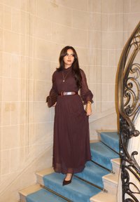 Długa, zwiewna sukienka w czekoladowo-brązowym kolorze z marszczonym taliowaniem, półprzezroczystą tkaniną i długimi rękawami. Dekoracyjny pasek i kopertówka, stojące na niebieskich schodach.