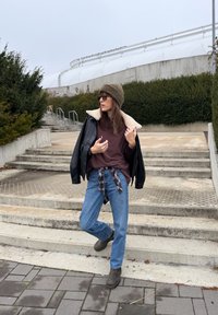 Crna kožna jakna s ovratnikom od ovčje kože, smeđa majica, plave traperice, smeđe čizme, zelena pletena kapa i sunčane naočale. Kockasta košulja vezana oko struka.