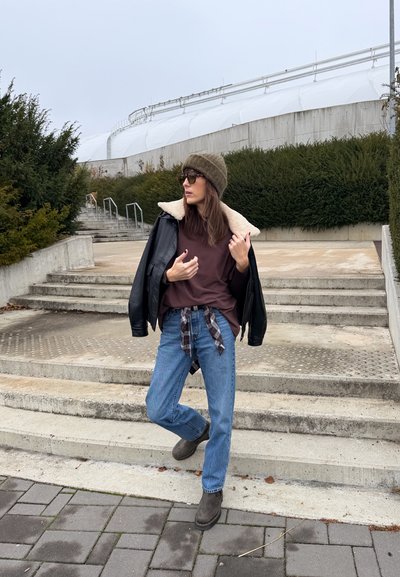 Chaqueta de cuero negra con cuello de borrego, camiseta marrón, vaqueros azules, botas marrones, gorro de punto verde y gafas de sol. Camisa de cuadros atada a la cintura.