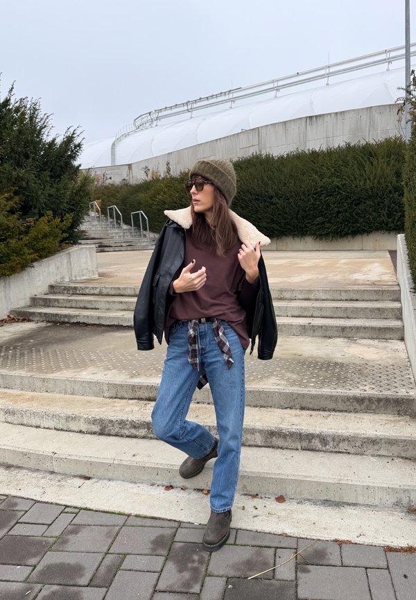 Black leather jacket with shearling collar, brown T-shirt, blue jeans, brown boots, green knitted beanie, and sunglasses. Checked shirt tied at waist.