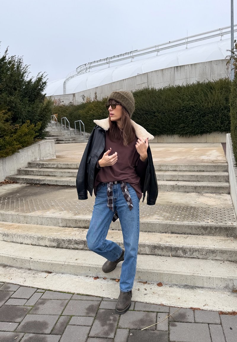 Must nahast jakk talje juures karvase kraega, pruun t-särk, sinised teksad, pruunid saapad, roheline kootud müts ja päikeseprillid. Talje juures seotud ruuduline särk.