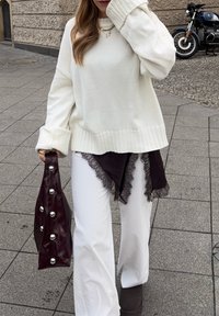 Bijeli oversized džemper s rebrastim manžetama, slojevit s crnim topom ukrašenim čipkom i širokim bijelim hlačama. Drži sjajnu bordo torbu ukrašenu srebrnim gumbima.