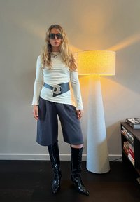 Femme aux longs cheveux blonds portant des lunettes de soleil surdimensionnées, un haut blanc à manches longues, une large ceinture noire, un short gris à hauteur des genoux et des bottes noires montant jusqu'aux genoux.