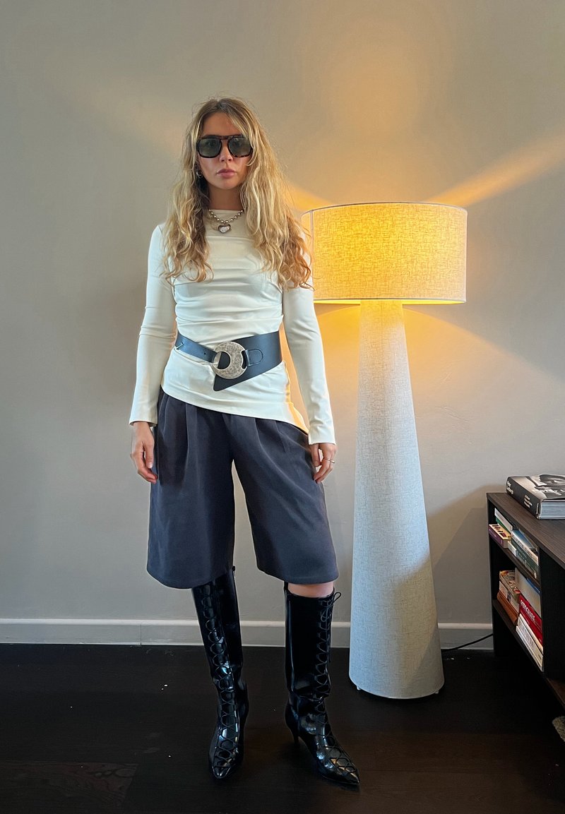 Femme aux longs cheveux blonds porte des lunettes de soleil surdimensionnées, un haut blanc à manches longues, une large ceinture noire, un short gris arrivant aux genoux et des bottes noires montantes jusqu'aux genoux.