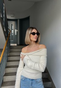 Bela pulover z odkrito ramo iz mehke in teksturirane pletenine, v kombinaciji s svetlo modrimi kavbojkami. Temna sončna očala dopolnjujejo videz. V ozadju so stopnice.
