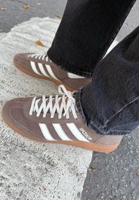 Brązowe zamszowe buty sportowe z białymi paskami, brązową gumową podeszwą i białymi sznurowadłami. Buty zostały zestawione z czarnymi dżinsami na teksturowanej powierzchni.