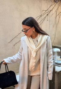 Manteau gris clair par-dessus un pull en maille torsadée crème, écharpe ivoire avec boutons décoratifs, et pantalon beige ; tenant un sac noir.