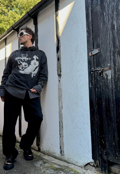 Sudadera negra con diseño gráfico, puños de camisa a rayas, pantalones negros y zapatos negros gruesos. El fondo presenta una pared blanca texturizada y una puerta de madera.