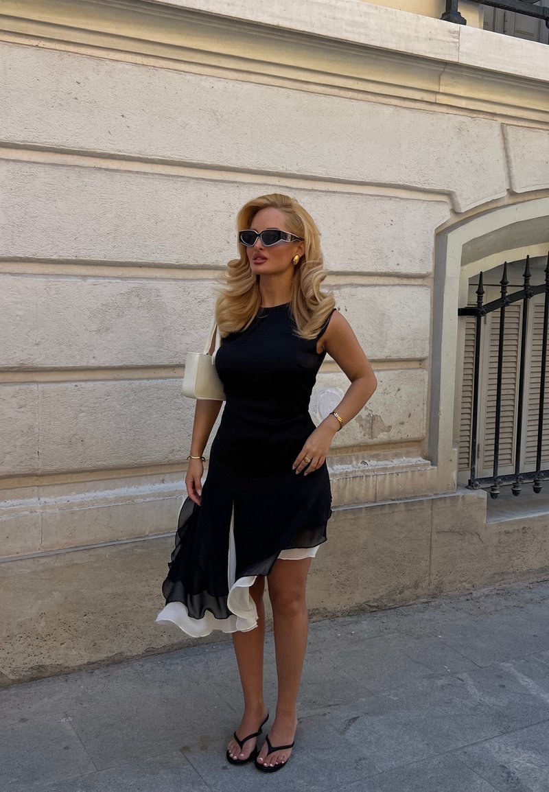 Γυναίκα με μακριά ξανθά μαλλιά, φορά μαύρο και άσπρο φόρεμα χωρίς μανίκια, μαύρα σανδάλια, γυαλιά ηλίου και κρατά μια λευκή τσάντα σε δρόμο της πόλης.