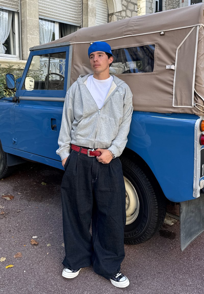Grigia felpa con zip, t-shirt bianca, pantaloni di jeans scuri larghi, cintura rossa e sneakers nere. Veicolo blu con cappotta beige sullo sfondo.