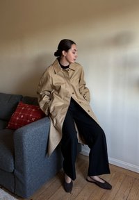 Beige, oversized trench coat z hladké tkaniny s velkým límcem a knoflíky, nošený přes černé kalhoty s širokými nohavicemi a tmavou obuv, sedící na šedé pohovce.