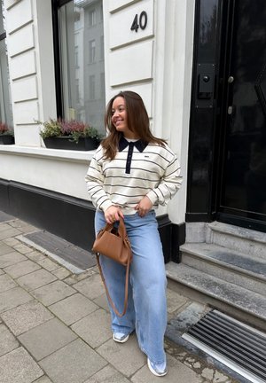 Mujer con suéter a rayas y jeans anchos sosteniendo un bolso marrón, de pie en la acera junto a un edificio blanco con una puerta negra con el número 40.