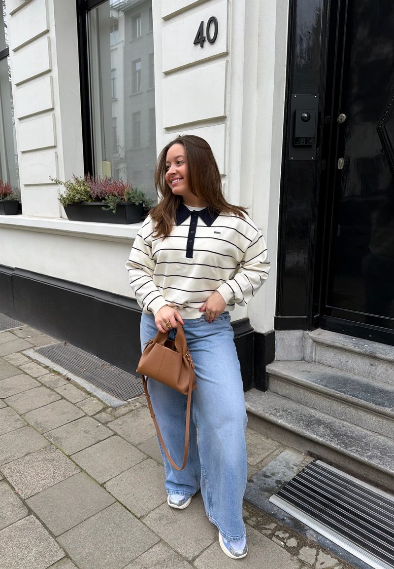 Femme en pull à rayures et jean large tenant un sac à main marron, debout sur le trottoir près d'un bâtiment blanc avec une porte noire numérotée 40.
