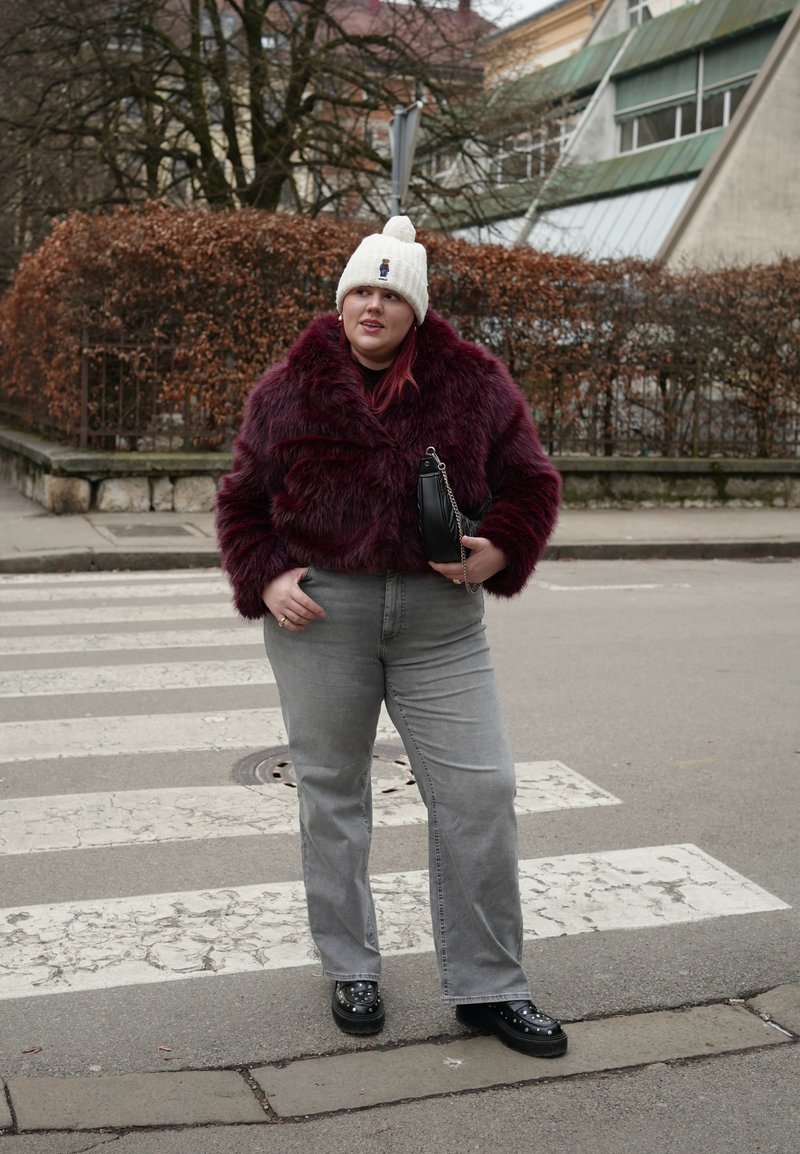 Ženska, oblečena v bordo bundu iz umetnega krzna, belo pleteno kapo, sive jeans hlače in črne čevlje, stoji na mestnem prehodu za pešce ob brezlistnih grmih.