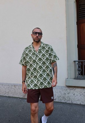 Camisa de manga corta en verde con un patrón floral, combinada con pantalones cortos marrones con acentos blancos. Gafas de sol y calcetines blancos completan el look.