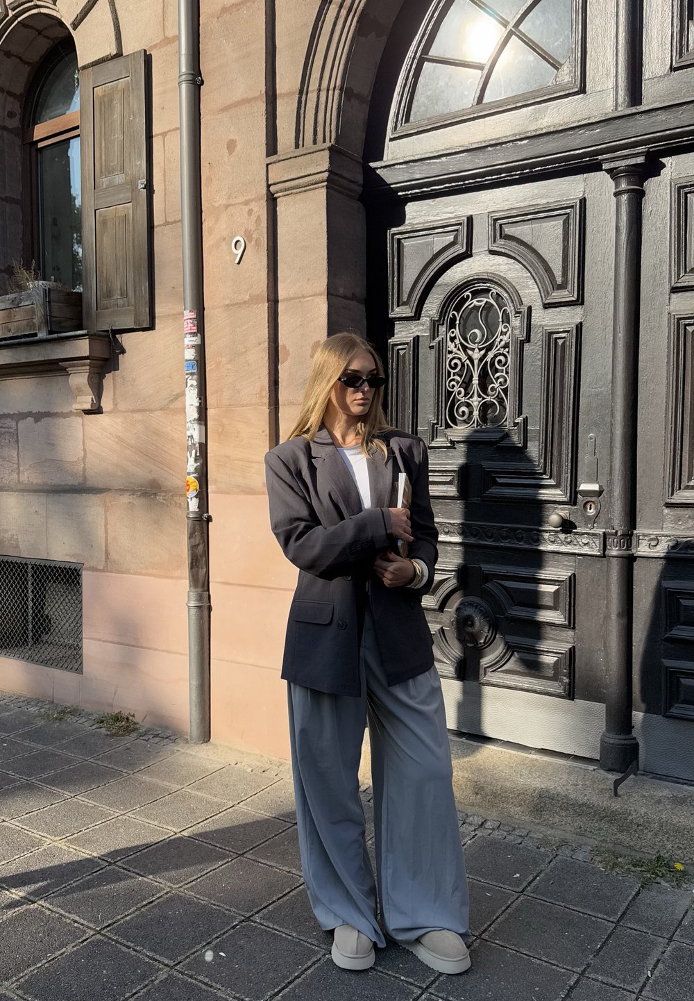 Przesadnie duża szara marynarka, biała bluzka i szerokie szare spodnie. Okrągłe beżowe buty i ciemne okulary przeciwsłoneczne dopełniają strój. W tle dekoracyjne drzwi.