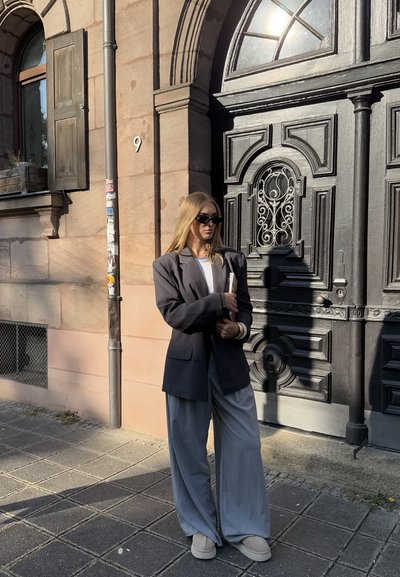 Blazer gris de gran tamaño, blusa blanca y pantalones grises de pierna ancha. Zapatos beige redondeados y gafas de sol oscuras completan el conjunto. Puerta decorativa al fondo.