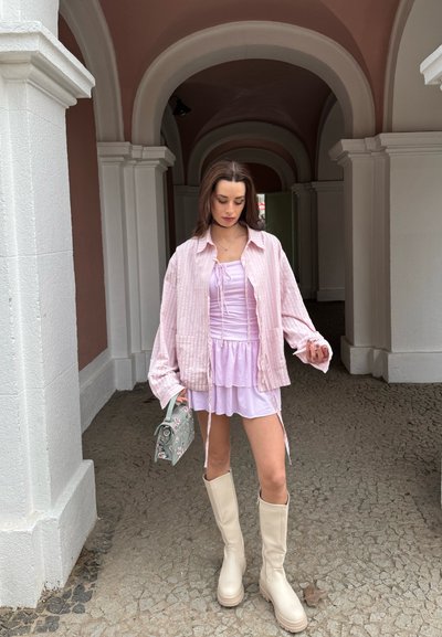 Mujer de pie bajo un arco, vistiendo un vestido lila claro, camisa rosa de gran tamaño, botas beige hasta la rodilla, sosteniendo un bolso floral verde.