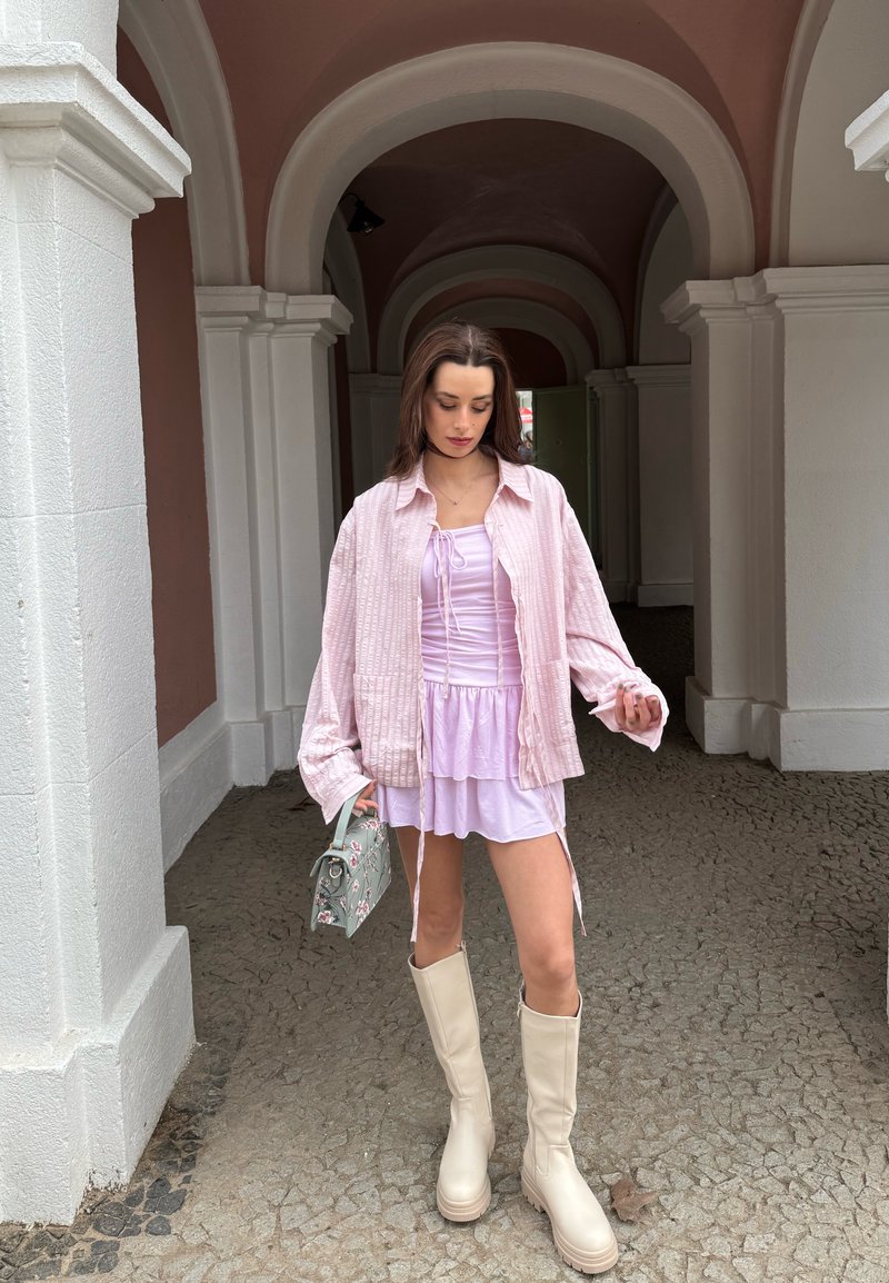 Woman standing under archway wearing light lilac dress, oversized pink shirt, beige knee-high boots, holding green floral handbag.