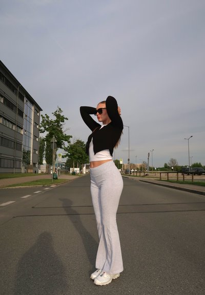 Pantalones de chándal grises acampanados, blusa blanca ajustada y cárdigan negro. Accesorios incluyen gafas de sol negras y zapatillas deportivas blancas. Entorno urbano.