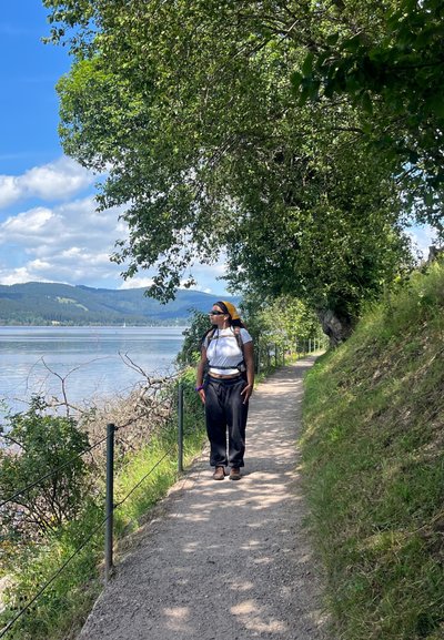 Persona de pie en un camino de grava junto a un lago, vistiendo una blusa blanca, pantalones negros y una cinta amarilla en la cabeza, rodeada de vegetación y montañas.
