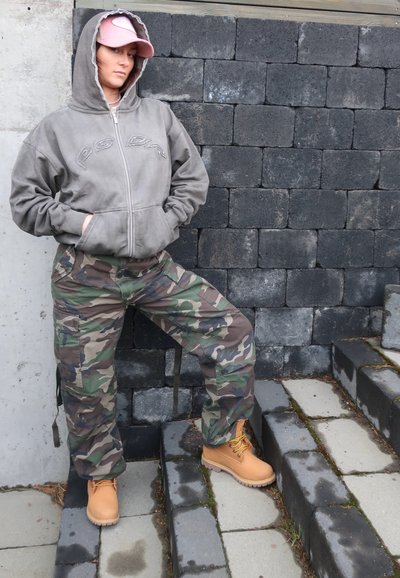 Sudadera gris con cremallera y logo bordado, pantalones cargo de camuflaje y botas de trabajo marrón con cordones amarillos, posando en unos escalones de piedra frente a una pared de bloques.
