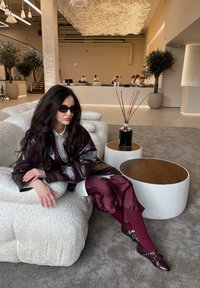Bruine leren jas, witte shirt, bordeauxrode broek en donkere schoenen. Geleefde, ronde bank in een moderne interieur met een verlichtingsarmatuur erboven.