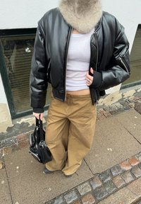 Personne portant une veste en cuir noire, un haut court blanc, un pantalon large beige, et portant un sac à main noir dans une rue pavée.