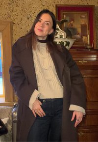 Γυναίκα με σκούρα μαλλιά, ντυμένη με κρεμ πουλόβερ πλεκτό με πλεξούδες και σκούρο παλτό, στέκεται μέσα σε εσωτερικό χώρο κοντά σε ξύλινα έπιπλα και έναν κορνιζαρισμένο πίνακα.