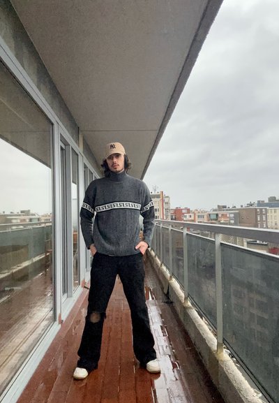 Jersey de cuello alto gris texturizado con patrón en contraste, combinado con pantalones negros y zapatillas blancas, llevado por una persona en un balcón.