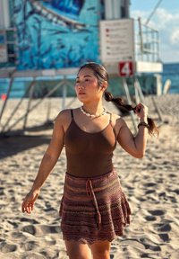 Jeune femme en haut marron et jupe à motifs tenant une tresse, debout sur une plage ensoleillée près d'une tour de surveillance et de l'océan.