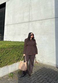 Brun skræddersyet blazer og matchende brede bukser lavet af blødt stof. Sorte højhælede støvler. Holder en struktureret beige håndtaske.