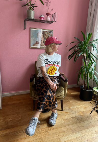 Camiseta gráfica con un diseño de pizza, combinada con una falda de estampado de leopardo, zapatillas gruesas grises y una gorra rosa, sentada en una silla vintage.