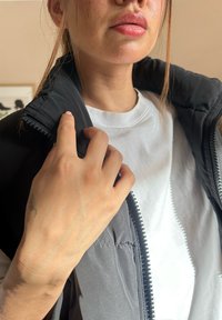 Juodas pūkinis liemenės su užtrauktuku ir aukštu apykakle, dėvimas ant baltos marškinėlio. Matoma ranka laiko apykaklę, rodanti odos toną ir tatuiruotę.