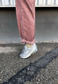 Ασημένια αθλητικά παπούτσια με υφή και γκρίζες λεπτομέρειες, φορεμένα με ρολά παντελόνια από ροζ κορδούρα. Το έδαφος είναι ανώμαλο και τραχύ.