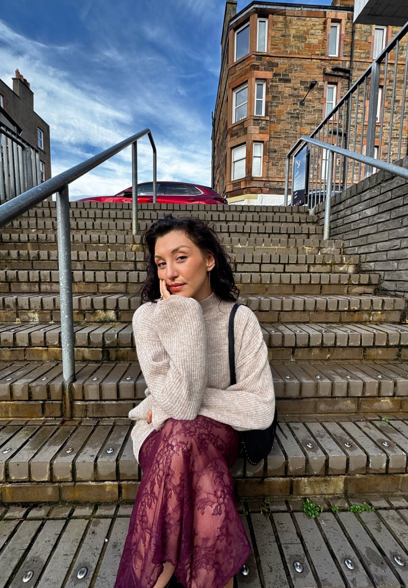 Knit beige sweater with oversized sleeves paired with a sheer, burgundy lace skirt. Subject seated on stone steps, natural texture visible.