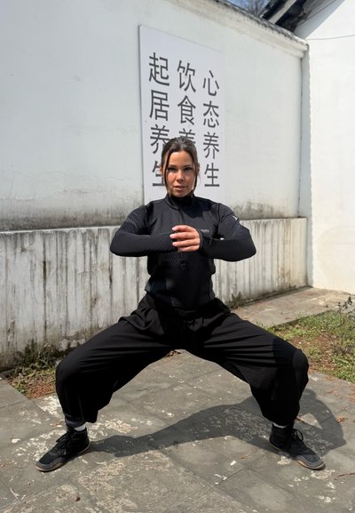 Mujer con ropa deportiva negra realizando una sentadilla amplia al aire libre cerca de una pared blanca con caracteres chinos.