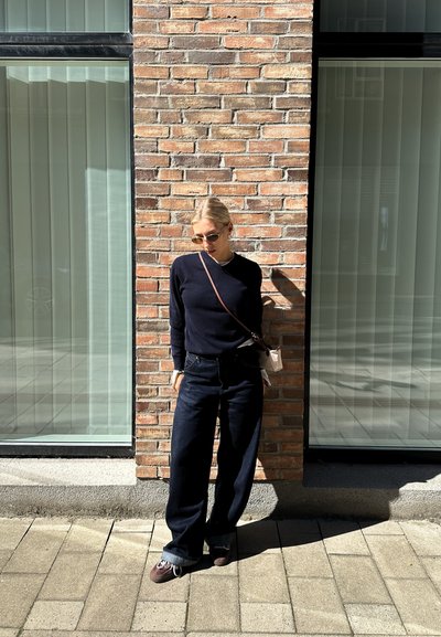 Suéter de punto azul marino, jeans oscuros de cintura alta y zapatillas marrones. De pie contra una pared de ladrillo con grandes ventanas de cristal.