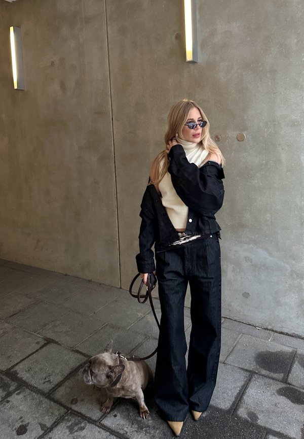 Femme en veste noire, col roulé beige et lunettes de soleil, debout sur le trottoir tenant une laisse avec un petit Bouledogue français.