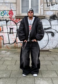 Une personne portant une doudoune noire par-dessus un t-shirt graphique, un jean noir ample avec une chaîne, et des baskets blanches, se tenant devant un graffiti.